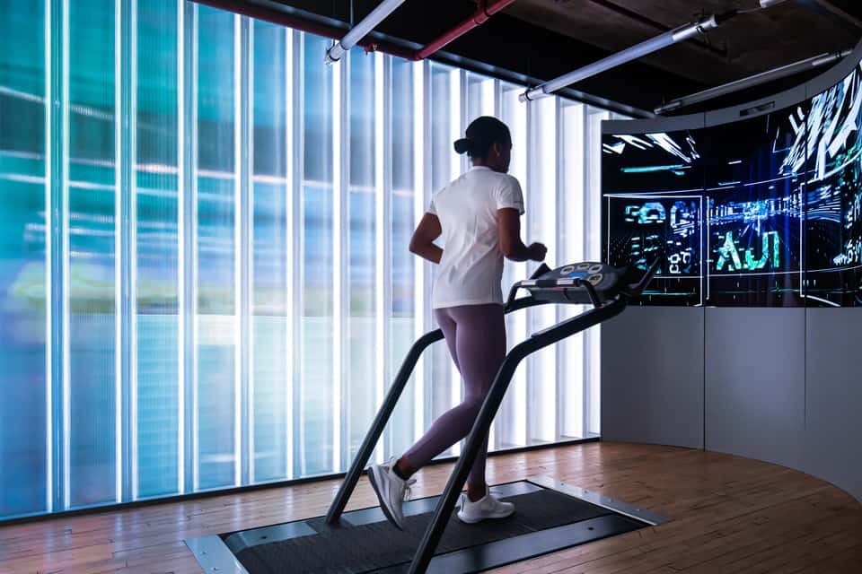 Photo of Adidas Running Lab installation in London showing a runner on a treadmill in front of a large LED screen displaying an abstract visualization of a cityscape.